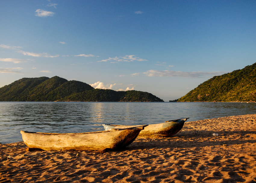 two carved wooden canoes sit on the sandy bank of Lake Malawi with green hills in the distance